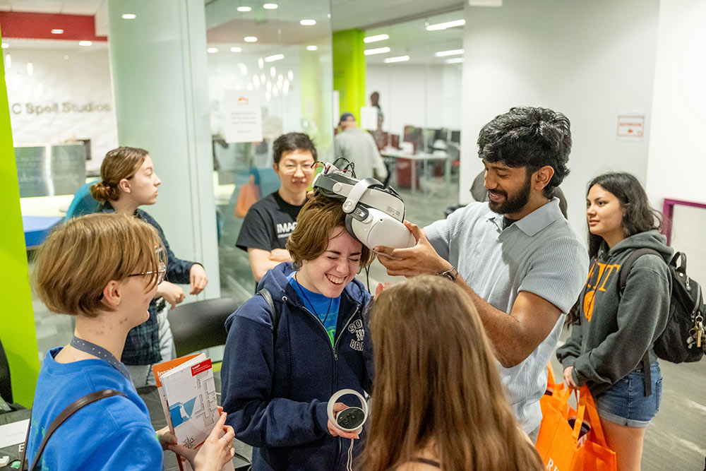 tudent places VR headset on Imagine RIT visitor while other visitors look  on.