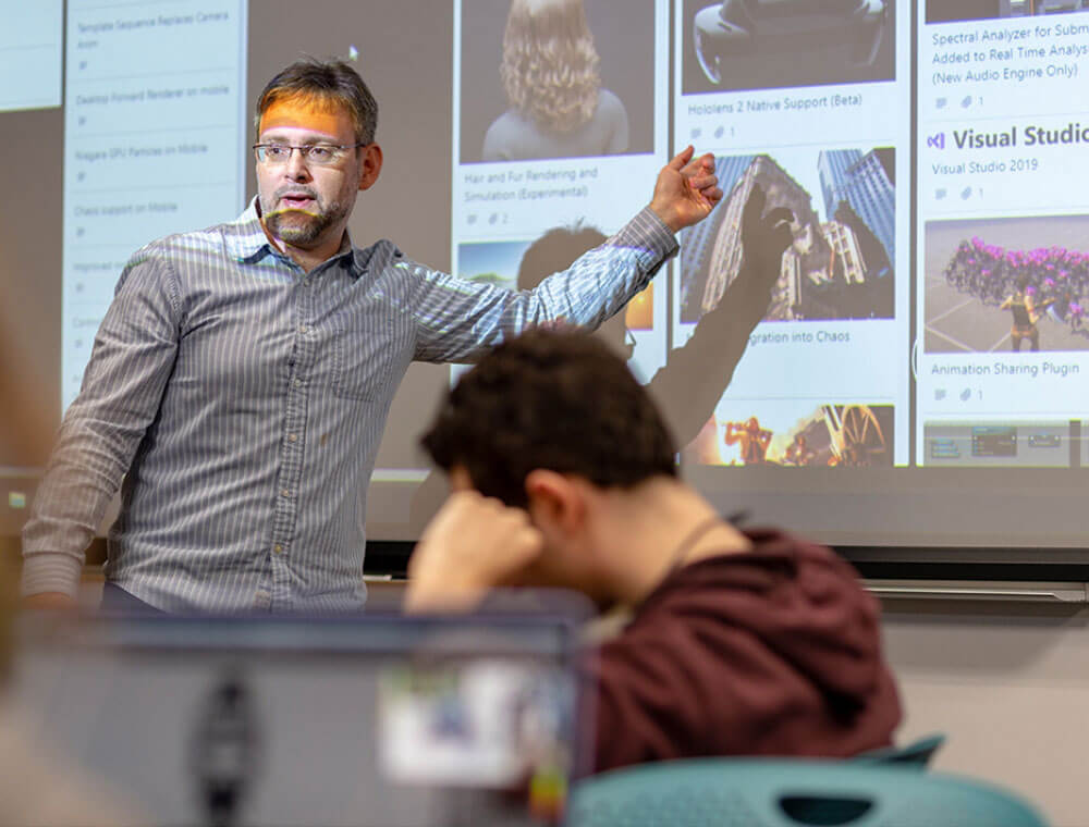 Shaun Foster stands in front of a presentation screen with a blog-like display of different animation projects. The light from the projector is cast over his face, and there is a student working at his desk in the foreground.
