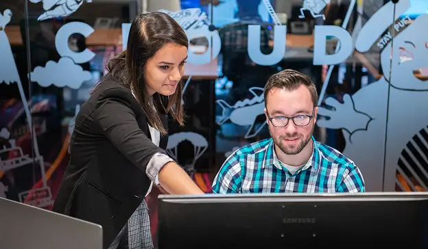 Two people at a workstation. One stands, pointing at a monitor, while the other sits and looks at the screen.