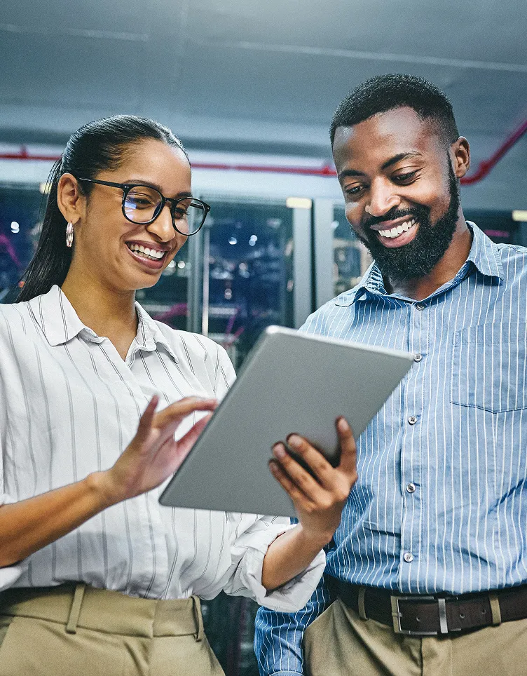 Two coworkers smile while reviewing something on a tablet in a work setting.