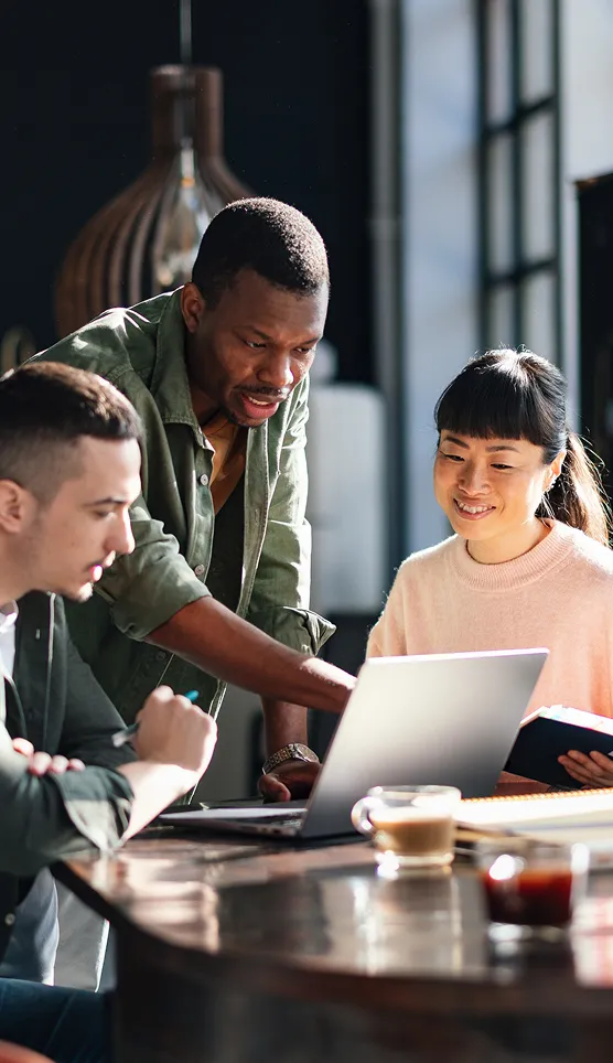 Three coworkers gather around a laptop in a bright office, with one person leaning in to point while the others look on.
