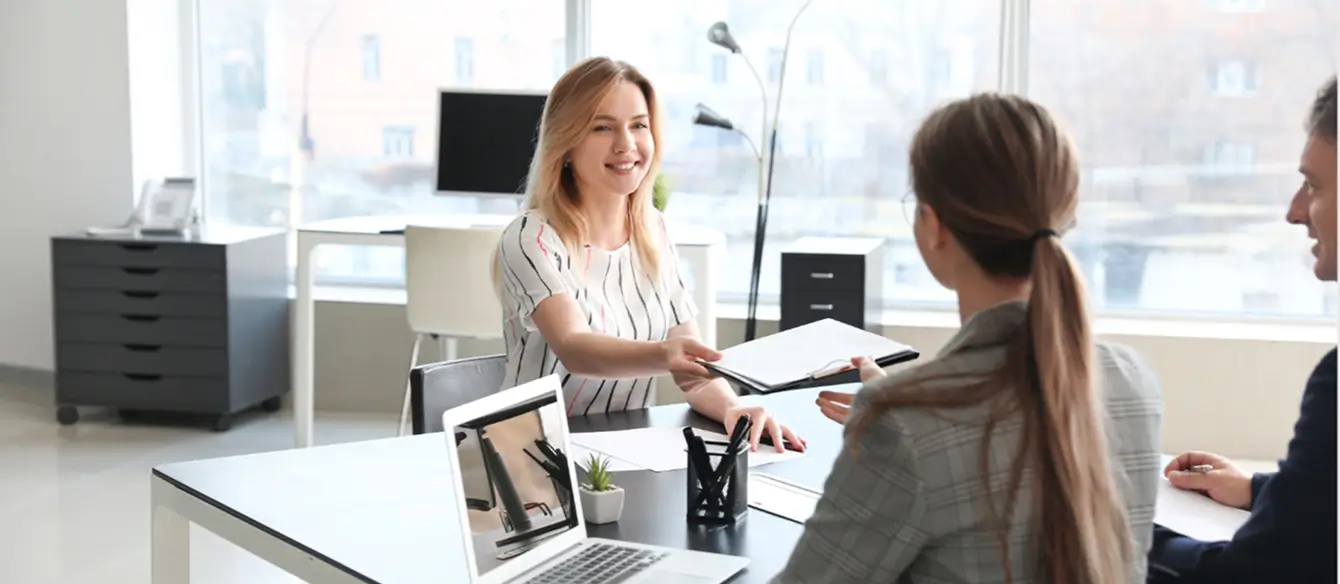 A person sitting behind a desk, handing a clipboard to two people sitting on the other side of the desk.