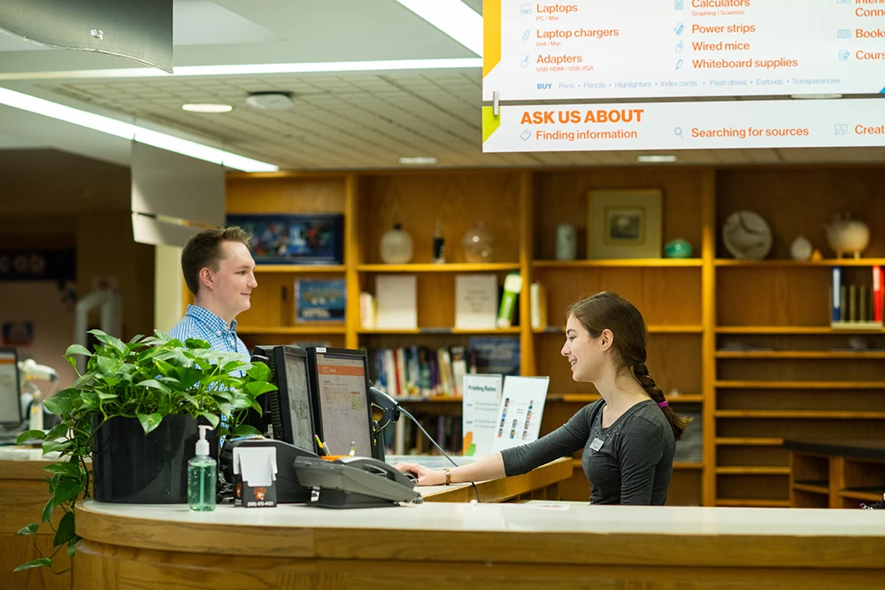 At a library service desk, an employee seated at a computer assists a visitor across the counter; bookshelves and an overhead sign listing resources are visible.