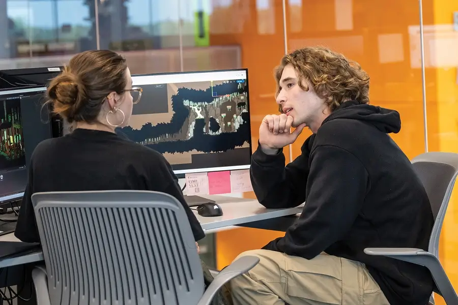 Two students sitting in front of a monitor showing a map in a game.