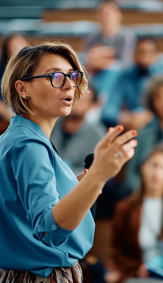 A person in glasses speaks into a handheld microphone and gestures while addressing an audience, with seated attendees blurred in the background.
