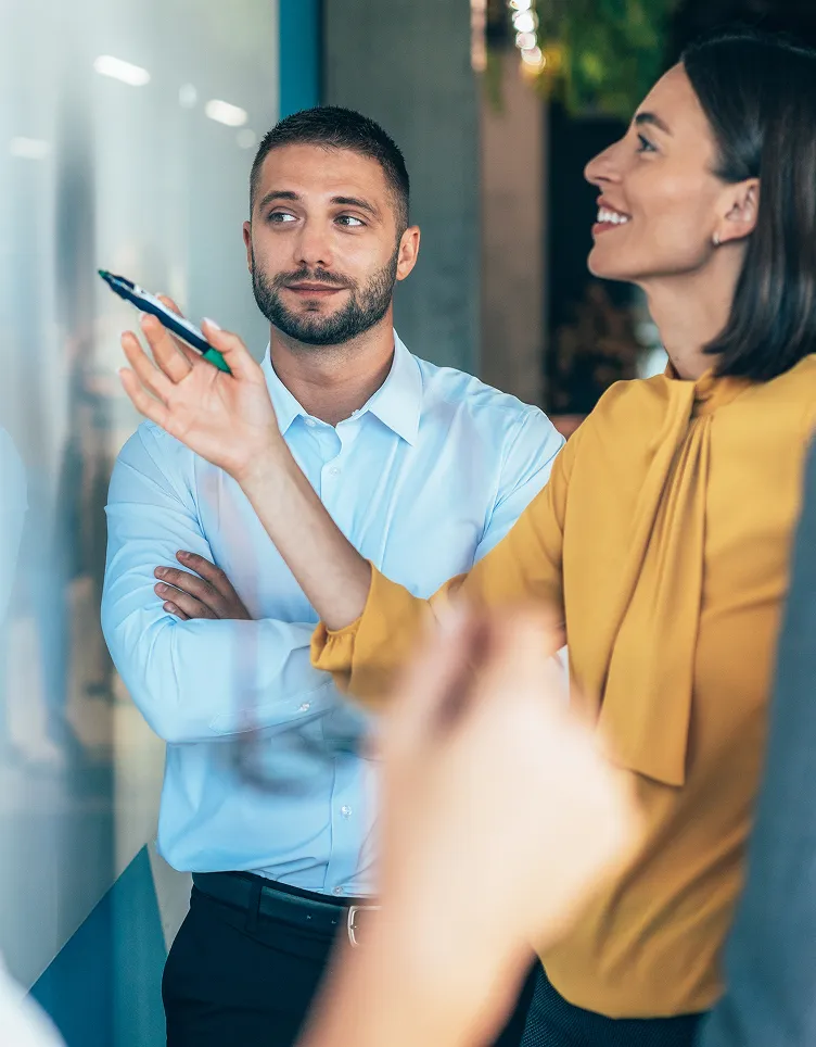 Two coworkers stand in an office discussion, with one person holding up a pen and the other listening with arms crossed.