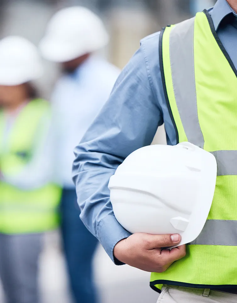 A person in a high-visibility safety vest holds a white hard hat at their side, with other hard-hat wearers blurred in the background.
