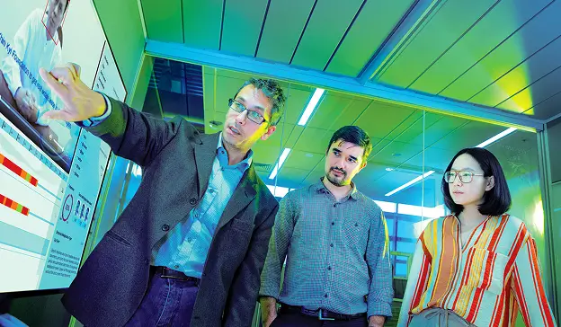 Three researchers standing in front of a large monitor, while one points to something on the screen.
