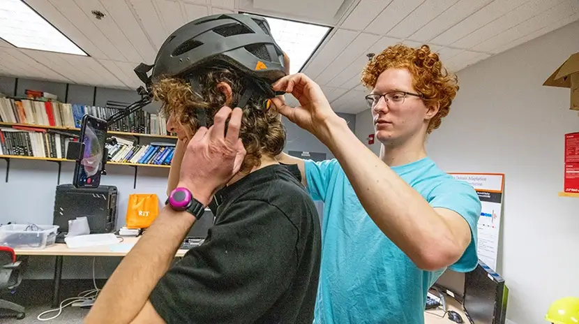 A person helping another put on a helmet that has a phone attached to it.