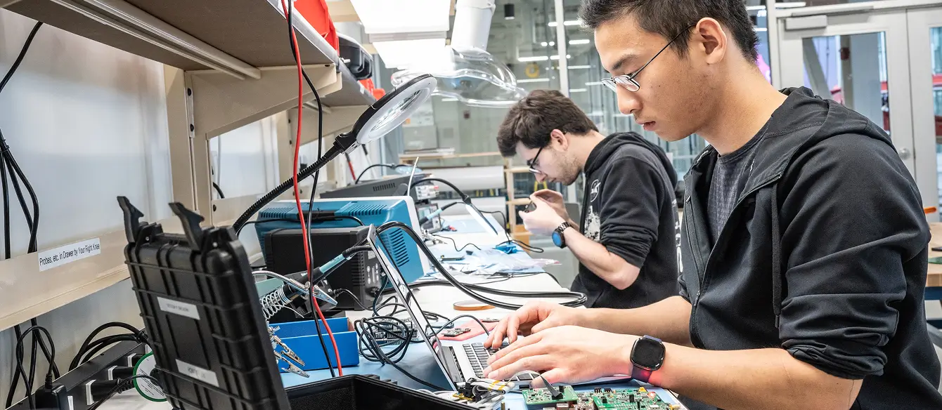 Two students working at laptops on a bench.