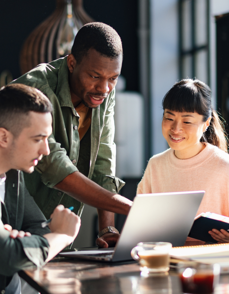 Three coworkers gather around a laptop in a bright office, with one person leaning in to point while the others look on.