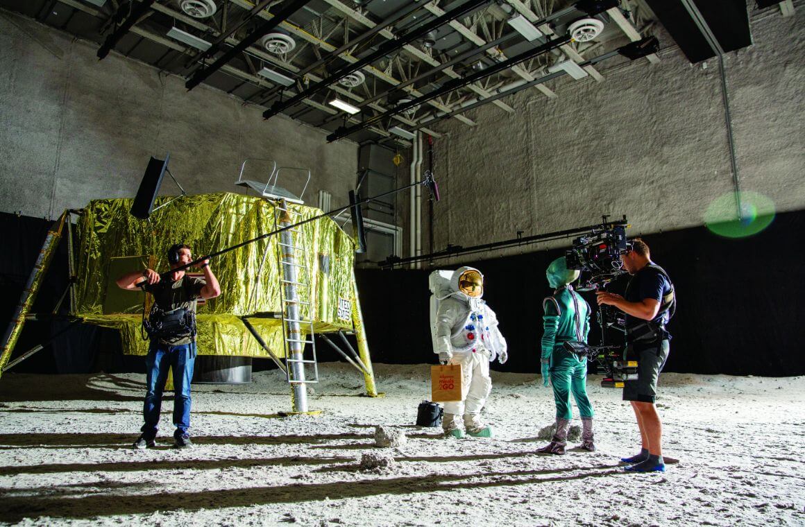 Two people dressed in costume as an alien and an astronaut while two others film and record audio on a set that looks like the moon.