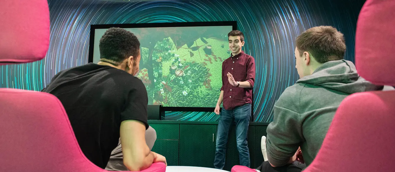 One person stands in front of a screen, displaying an environment from a video game, presenting to two people sitting in chairs.
