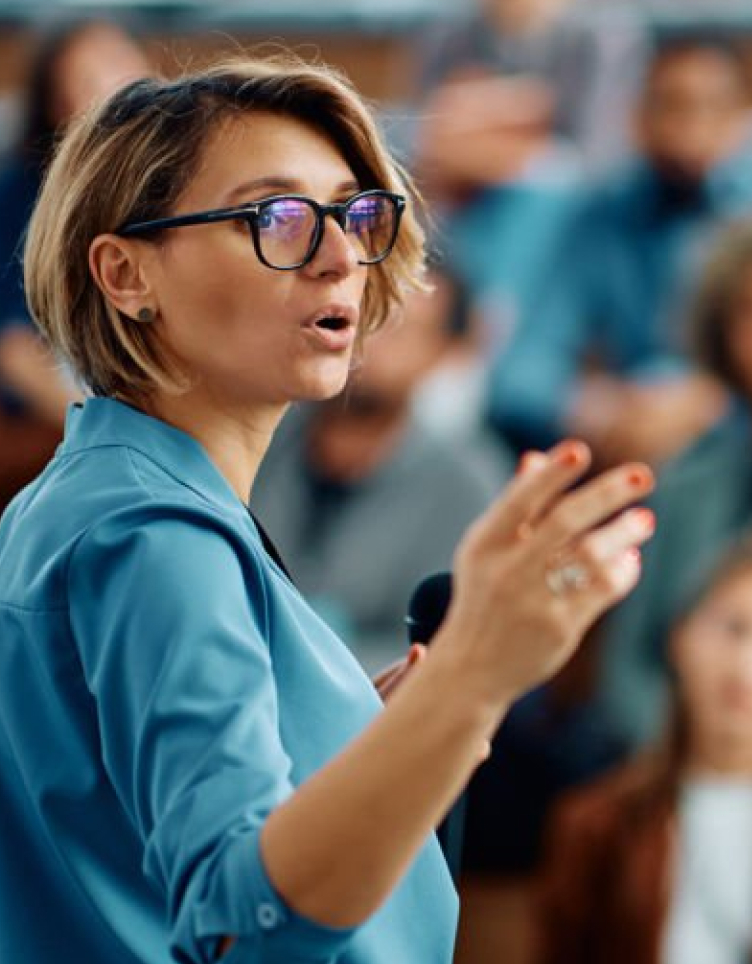 A person in glasses speaks into a handheld microphone and gestures while addressing an audience, with seated attendees blurred in the background.