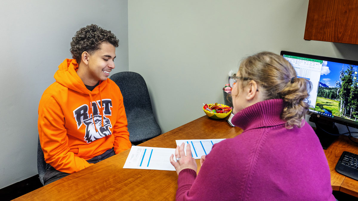 A student sits at an advisor's desk working with them.