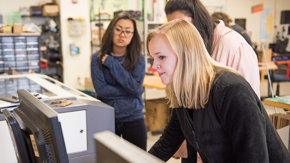 A student works on a computer next to a printer or laser cutter.