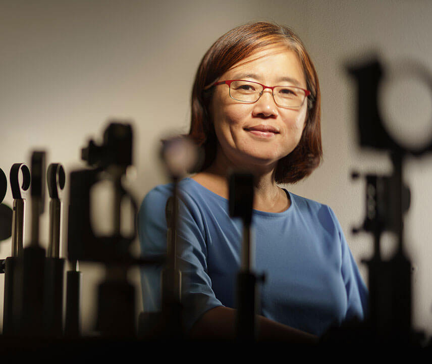 Associate professor Jie Qiao standing in the background with optical instruments in the foreground.
