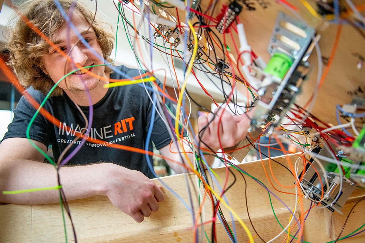 Man in Imagine RIT shirt looks at dozens of colorful wires hanging on the  underside of a pinball machine.
