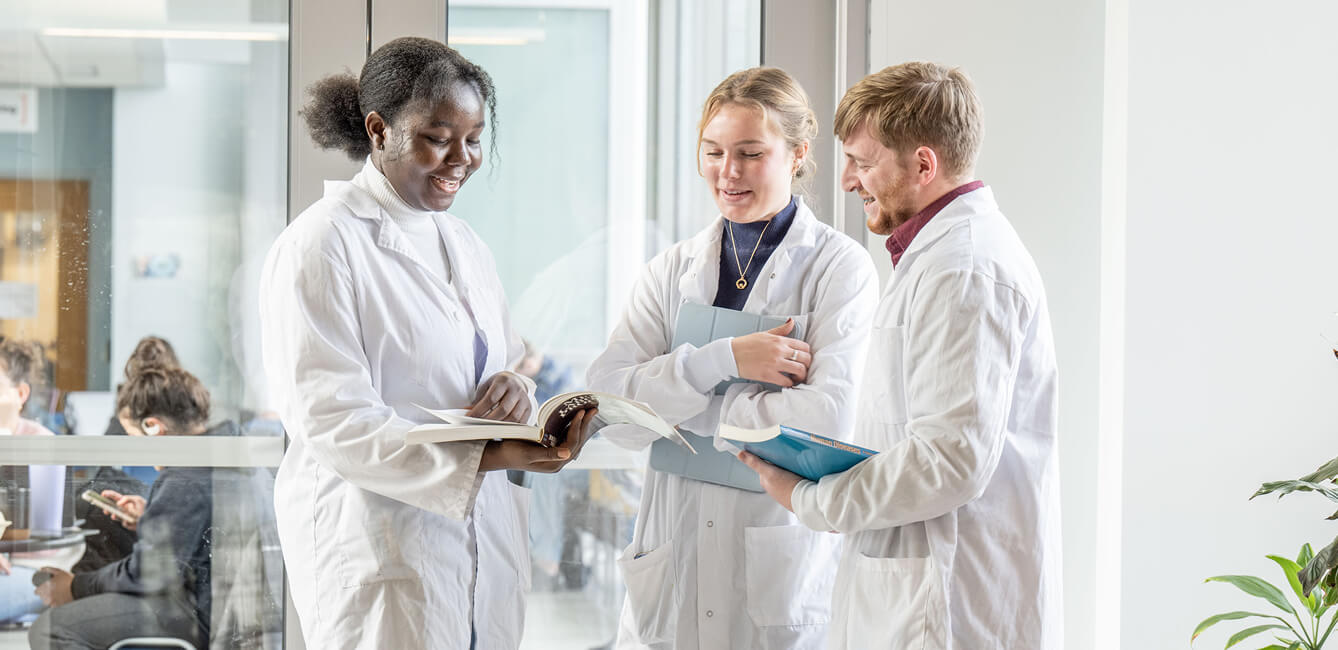Three people in lab coats, standing in a hallway, looking at an open medical book.