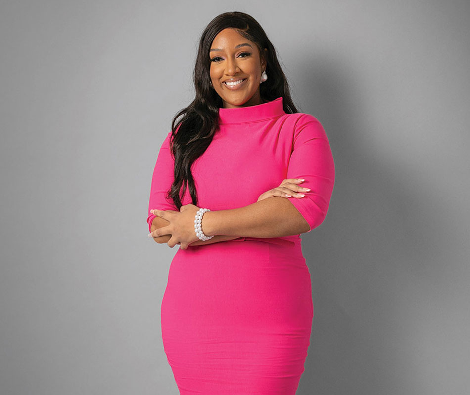 Shakierah Smith is shown standing in front of a gray wall wearing a hot pink dress with her hands crossed across her torso.