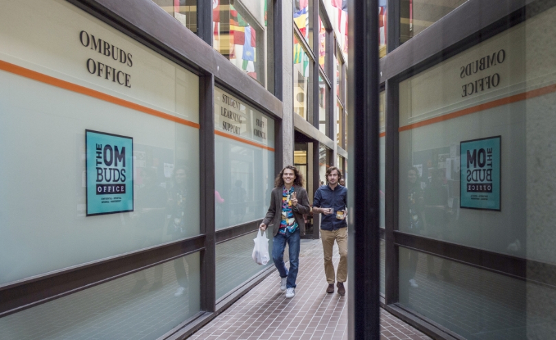 two people walking down a hallway with big windows with the ombuds logo on them