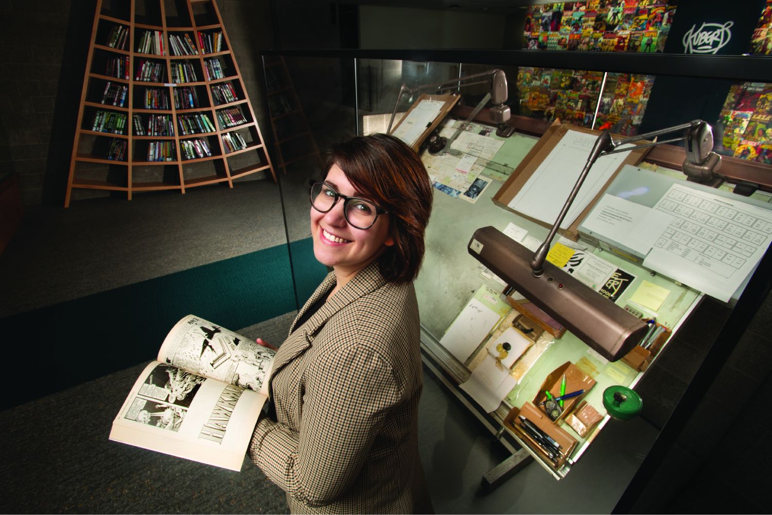 Leah Humenuck standing by a drafting table with a book open and looking over her shoulder