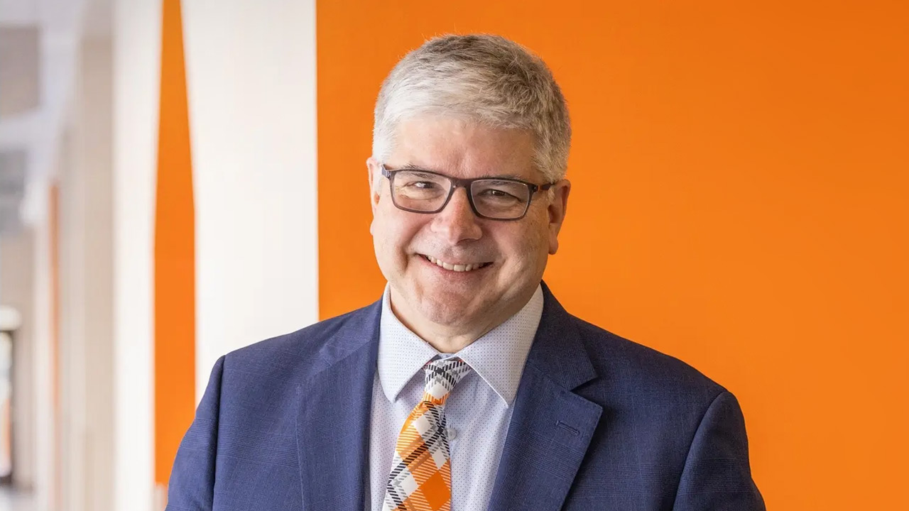 RIT President, Dr. William H. Sanders, smiling while standing in front of an orange wall.