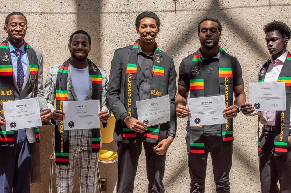 five Black college students wearing Black grads regalia and holding up certificates.