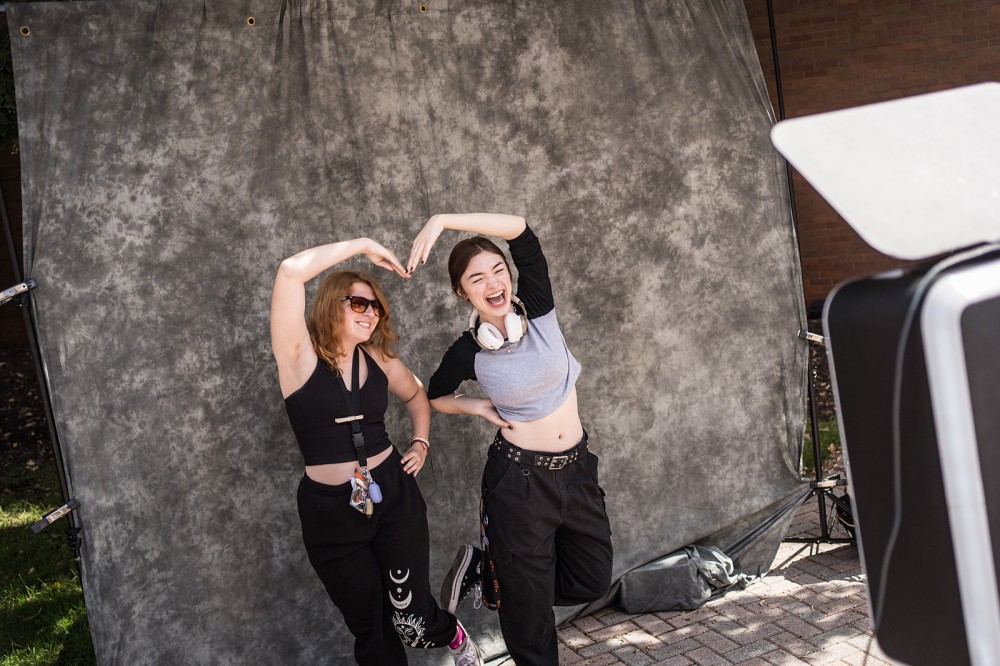 two college students posing for a photo against a gray backdrop, with their arms over their heads to form a heart.