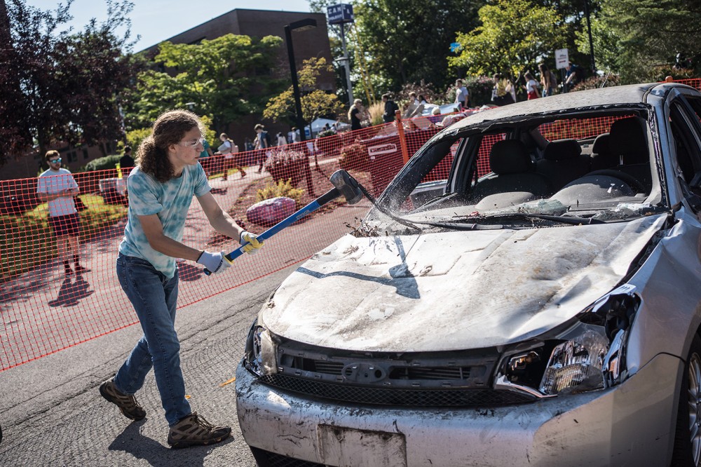 college student smashing the hood of a beat-up car with a sledgehammer.