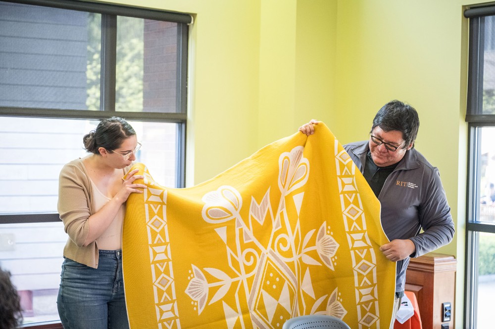 a younger woman and an older man hold up a yellow and white native american blanket.