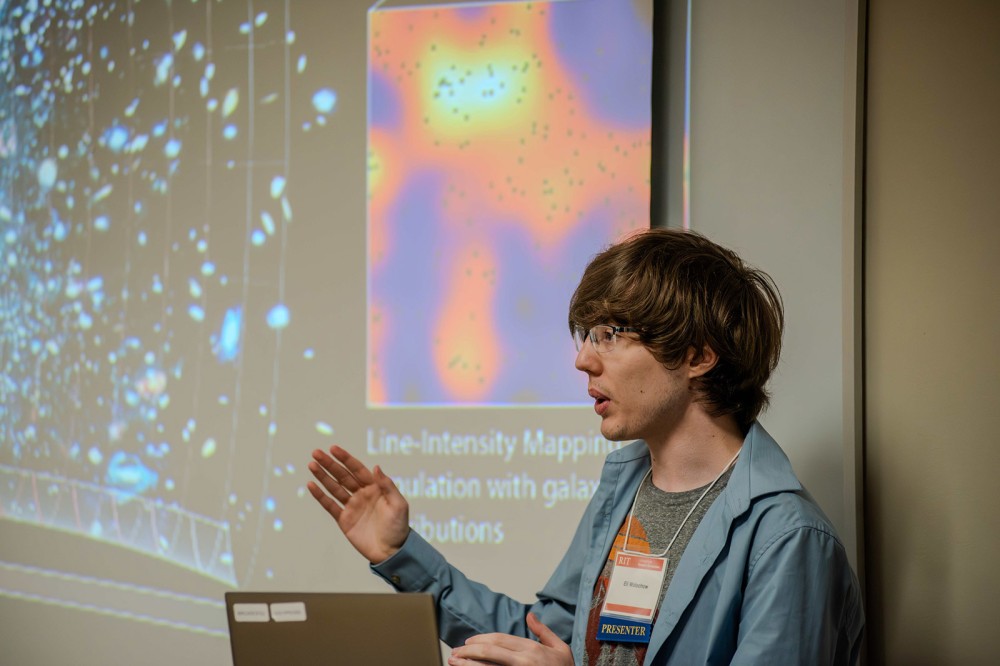 a student is shown standing in front of a presentation screen and he appears to be directing conversation to an audience.