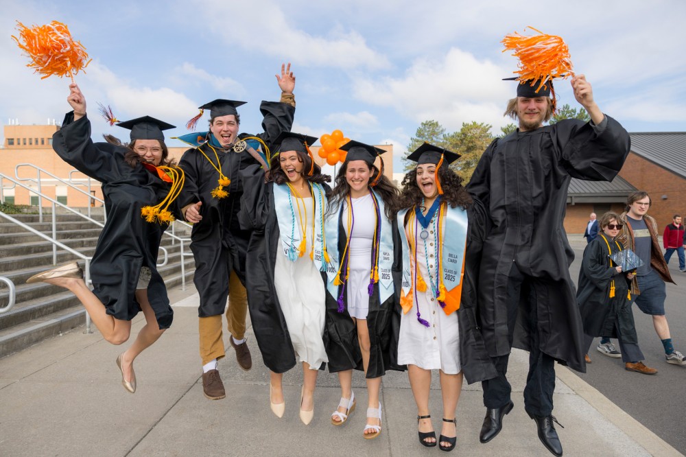 Graduates in caps and gowns cheer and jump on steps outside a campus building.