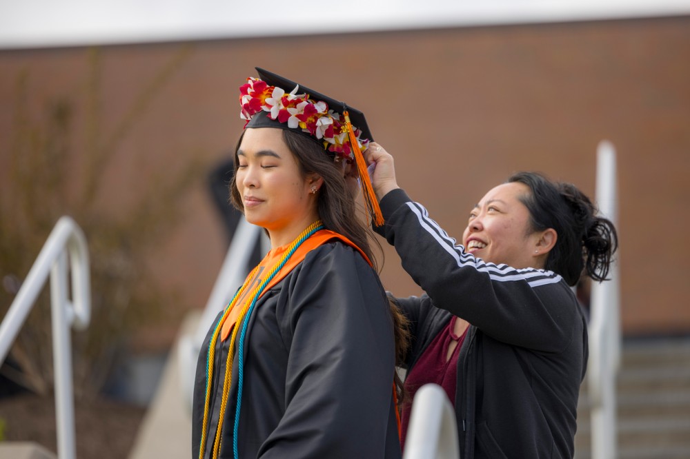 Woman helps adjust a floral crown on a smiling graduate in cap and gown.
