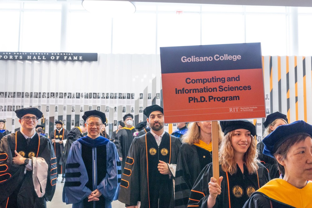 Graduates in academic regalia stand in a hallway under a Golisano College Computing and Information Sciences Ph.D. Program sign at RIT commencement.