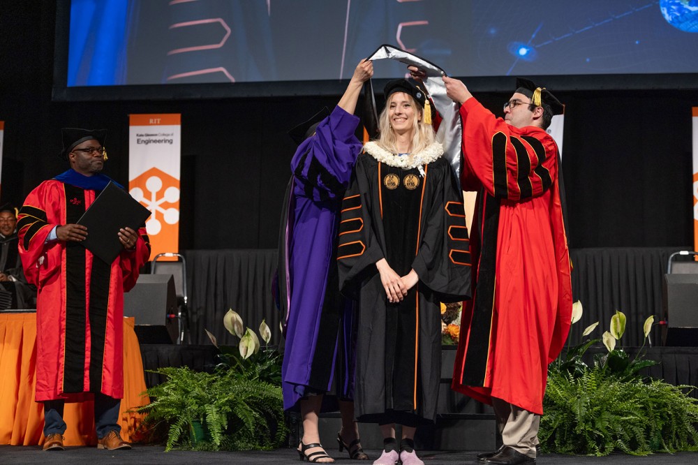 Faculty place a doctoral hood on a smiling graduate in academic regalia during an RIT commencement ceremony.