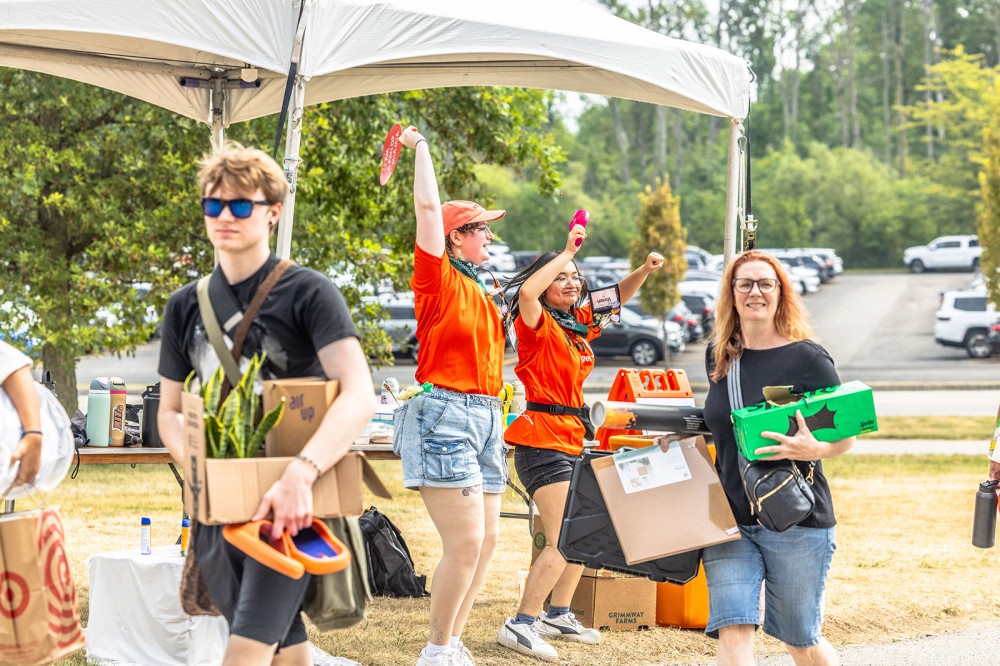 Students and volunteers carry boxes and supplies during college move-in under a tent.