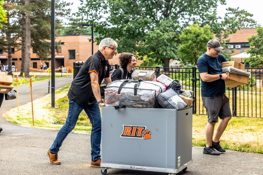 Man pushes a full cart with dorm supplies as others walk alongside on R I T campus.