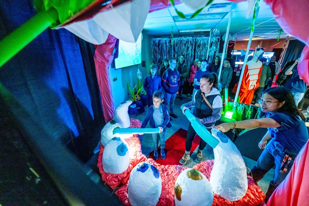 Visitors in a brightly lit, themed room watch and interact with a playful display featuring oversized teeth props and glowing blue foam “toothbrush.” A crowd stands in the background under colorful lighting and decorations