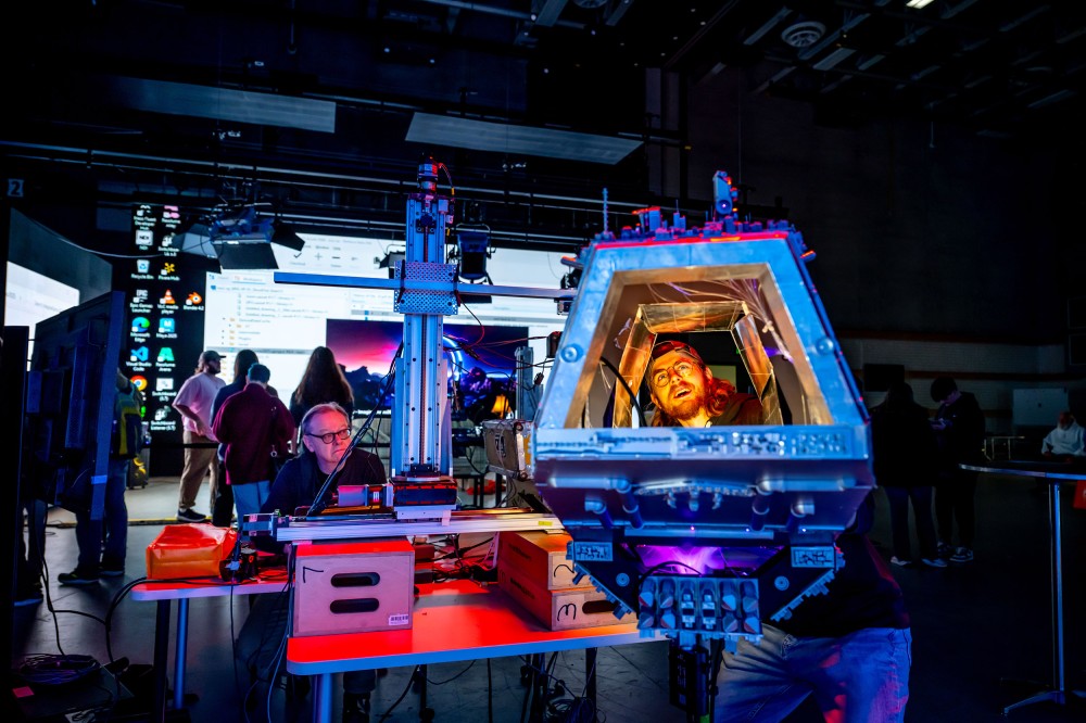 In a dark, neon-lit lab space, a student looks out from inside a large blue machine or cockpit-like frame on a worktable. People and a large projected computer screen are visible in the background.