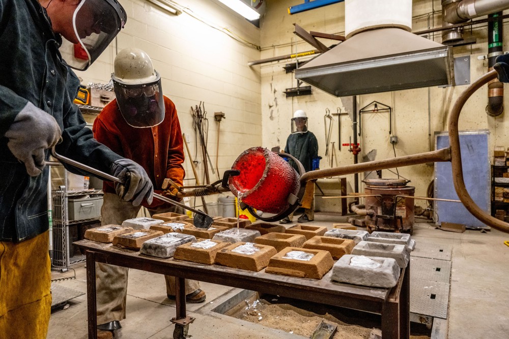 Two people in protective gear pour glowing molten metal from a crucible into molds lined up on a worktable. A workshop furnace and tools are visible in the background.