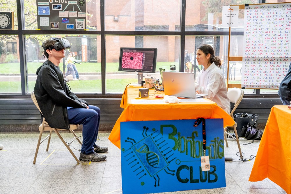 A student wearing a virtual reality headset sits at a bioinformatics club table while another student speaks across from a laptop and monitor showing a protein model. A sign reading “Bioinformatics Club” is displayed on the front of the table.