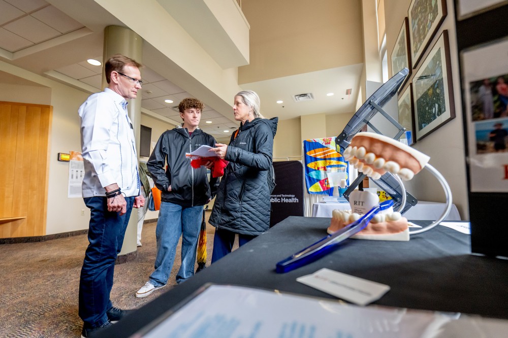 A prospective student and parent speak with an RIT staff member in a campus hallway near a table display with dental models. A sign for RIT’s global public health program is visible in the background.