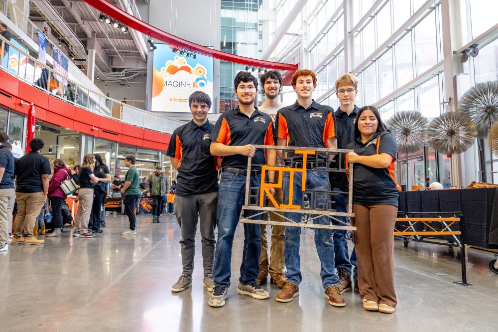 Six students in black-and-orange team shirts pose inside a large glass-walled atrium at an event, holding a metal display frame with orange “RIT” letters. Tables, attendees, and an “Imagine RIT” sign are visible in the background.