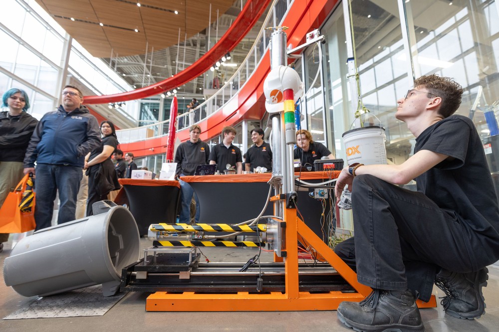 A student sits beside a large orange test rig with warning stripes and a vertical signal light while other students and visitors gather around tables in a bright atrium. A red rocket display is visible in the background.