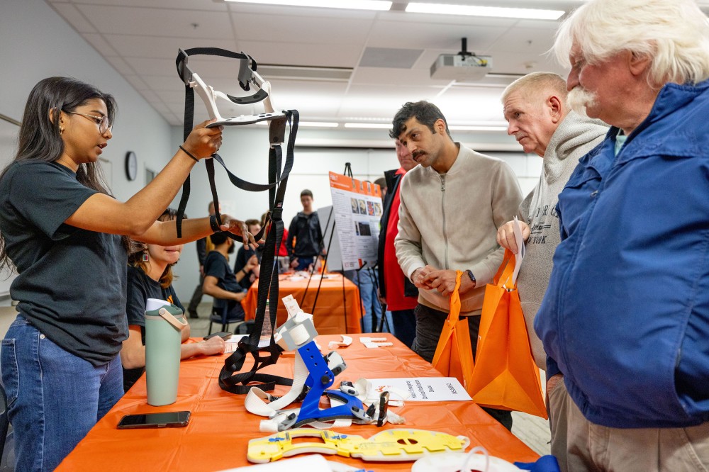 A student demonstrates a cervical collar or neck brace prototype at an event table while visitors watch and ask questions. Prototype parts and straps are spread across the table.