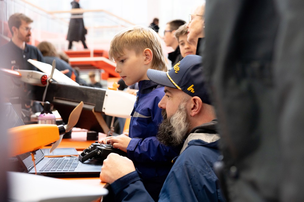 A child and an adult use a laptop and handheld controller to operate a model aircraft at a crowded indoor event. The plane’s propeller and part of the aircraft body are visible in the foreground.