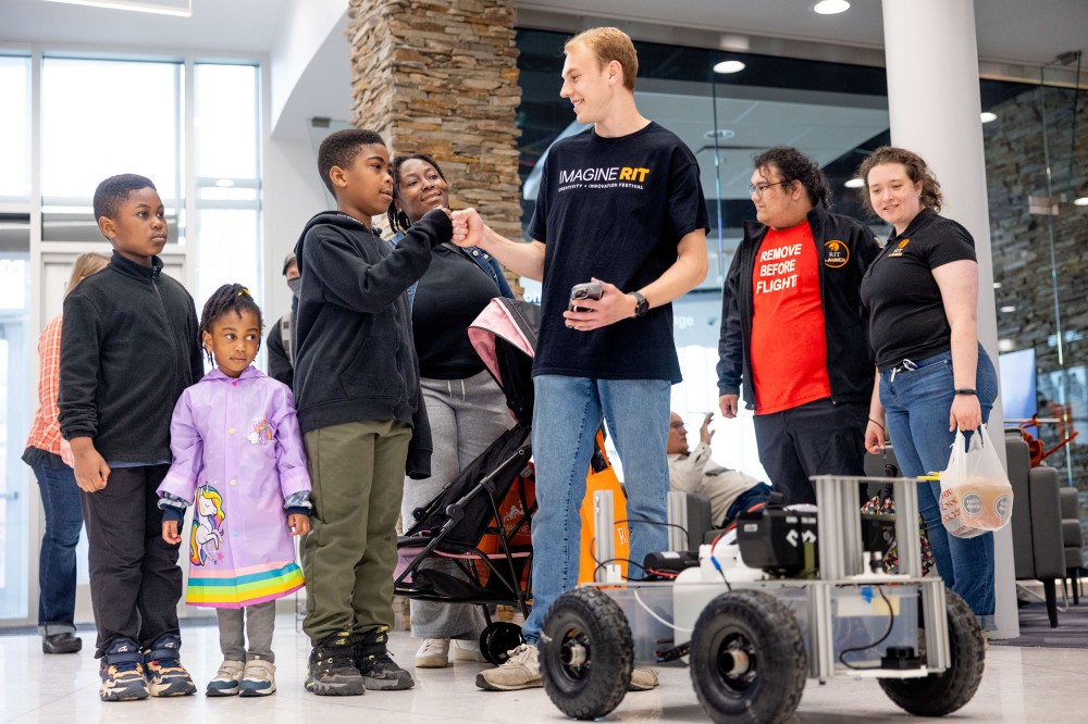 A student demonstrates an autonomous wheeled robot to visitors, including children, in a bright indoor atrium. The robot sits in the foreground with sensors and wiring visible as the group watches.