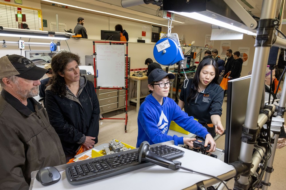 A student facilitator guides a participant through a robotics challenge at a workstation in the Toyota Lab while visitors watch. The table holds tools and computer equipment, with other lab stations and attendees in the background.