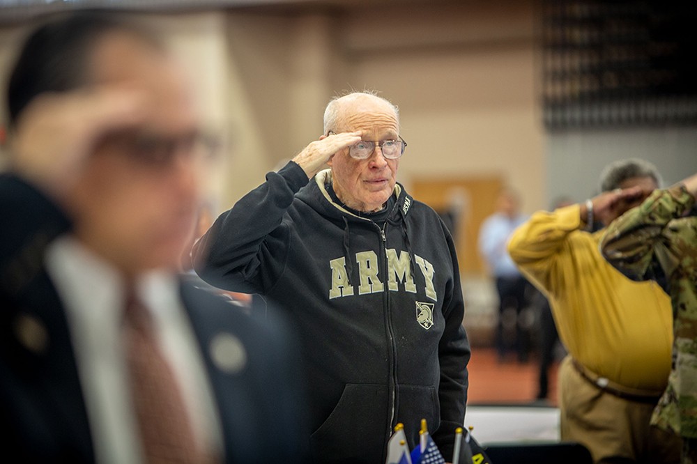 A crowd of people saluting including a person with a sweatshirt that says ARMY.